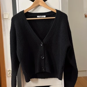 Svart kofta från NA-KD, stickad - Mysig svart stickad kofta från NA-KD med v-ringning och tre stora knappar framtill. Koftan har lång ärm och en lite croppad passform som ger en trendig look. Tröjan är inte sticksig!