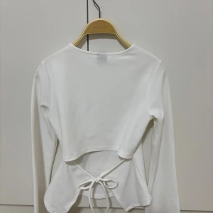 Vit långärmad topp med knyt från Gina Tricot - Säljer en stilren vit långärmad topp från Gina Tricot i storlek XS. Toppen har en rund halsringning och snygg knytning i midjan baktill som ger en extra detalj. 