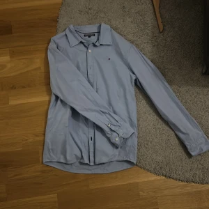 Ljusblå skjorta Tommy Hilfiger 164 - Klassisk ljusblå skjorta från Tommy Hilfiger med liten broderad logga på bröstet. Skjortan har lång ärm, knappar framtill och är tillverkad i mjuk bomull. Perfekt för en clean och stilren look.
