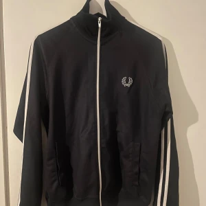  Fred Perry zip - Svart track jacket från Fred Perry med vita ränder längs ärmarna och vit dragkedja. Klassisk logga broderad på bröstet. Jackan har hög krage, två fickor och är tillverkad i mjuk polyester. Perfekt för en sportig och stilren look. Pris kan diskuteras 