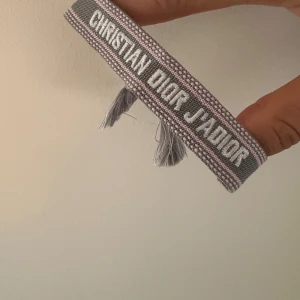 Amband grå/rosa - Snyggt vävt armband med texten 'J'Adior' i vitt. Armbandet är grått med rosa detaljer och har justerbara tofsar i ändarna. Perfekt accessoar för att lyfta din outfit med en exklusiv touch.