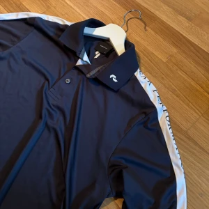 Blå långärmad pikétröja Peak Performance - Snygg mörkblå långärmad pikétröja från Peak Performance med vit rand längs ärmarna och logga på kragen. Tröjan har klassisk krage, knäppning framtill och är tillverkad i ett mjukt, sportigt polyestermaterial. Perfekt för en golfrunda.