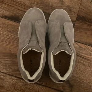 Grå mockasneakers från Eytys, stl 39 - Säljer ett par stilrena grå sneakers från Eytys i mocka med vit sula och diskret lila detalj bak. Skorna har elastiska paneler istället för snörning och en klassisk, rund tå. Perfekta för dig som gillar minimalistisk design och hög komfort.