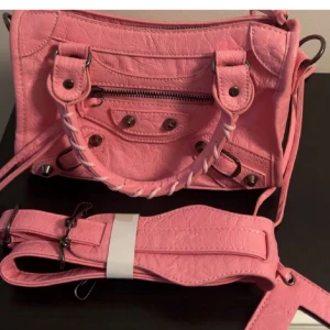 Rosa axelväska med flätat handtag - Cool rosa axelväska med snygga metalldetaljer och flätat handtag. Väskan har dragkedja framtill och kommer med en bred axelrem. Perfekt för dig som vill sticka ut med en färgglad accessoar.