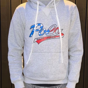 Grå hoodie Polo Ralph Lauren - Snygg grå hoodie från Polo Ralph Lauren med stor logga framtill i blått, vitt och rött med stjärnmönster. Tröjan har huva med vita snören och en klassisk känguruficka. Perfekt för en chill och sportig look.