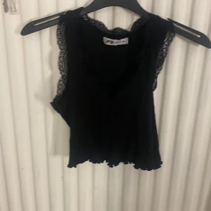 Svart linnetopp med spetsdetaljer - Snygg svart linnetopp från F2 med v-ringning och spets längs kanterna. Toppen är ärmlös och har en lätt vågig nederkant som ger en cool touch. Perfekt att styla med jeans eller kjol för en trendig look.