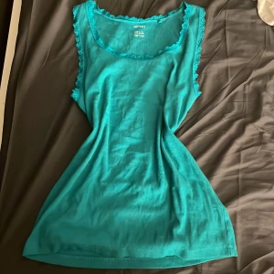 Turkos linnetopp med spets från Esmara - Snygg turkos linnetopp från Esmara i storlek XL. Toppen har rundad halsringning och ärmlös design med fina spetsdetaljer längs hals och ärmkanter. Mjuk och stretchig bomull som känns skön mot huden. Perfekt för dig som gillar färg och enkel stil.