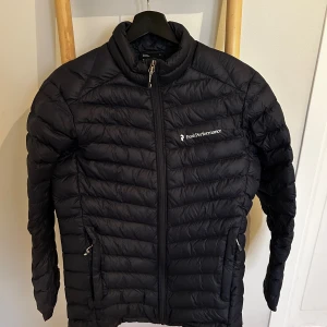 Svart dunjacka Peak Performance S - Svart dunjacka från Peak Performance i storlek S. Jackan har quiltad design, dragkedja framtill och två sidofickor med dragkedja. Märket syns på bröstet och ärmen. Perfekt för kalla dagar och har en stilren look som funkar till det mesta. Jackan har ett litet hål på ena ärmen som syns på sista bilden.