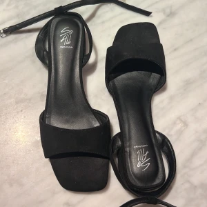 Svarta sandaler med låg klack So All - Stilrena svarta sandaler från So All med fyrkantig tå, bred rem över foten och smal ankelrem med spänne. Liten i storlek.     Skorna har låg klack och platt sula, tillverkade i syntetmaterial med mockakänsla. Perfekta för dig som gillar minimalistisk och trendig look. 