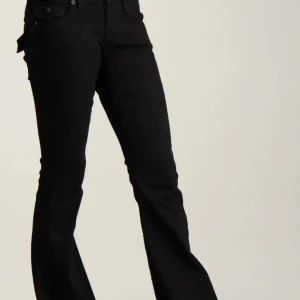 Svarta bootcut jeans med fickdetaljer - Svarta jeans från True Religion med bootcut-passform och snygga fickdetaljer bak. Jeansen har klassisk femficksdesign, låg midja och är tillverkade i stretchig denim för en bekväm känsla. Perfekta för dig som gillar en stilren och trendig look.