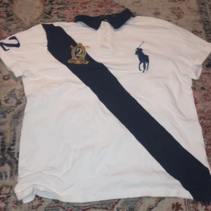 Vit pikétröja från Ralph Lauren - Vit pikétröja från Ralph Lauren med marinblå krage och en bred diagonal marinblå rand över framsidan. Stort broderat polospelare-märke på bröstet och broderad siffra på ärmen. Klassisk piké med knappar vid halsen.