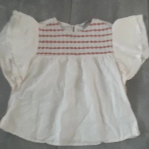 Vit blus med röd mönstrad överdel LCW - Supersöt vit blus från LCW Girls med vida korta ärmar och rund hals. Överdelen har ett rött, randigt mönster i broderad stil som ger en unik look. Blusen är luftig och har en lätt, flowy passform – perfekt för vår och sommar. Xxs/xs!