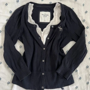 Mörkblå vintage kofta från Abercrombie & Fitch - Mysig vintage mörkblå kofta från Abercrombie & Fitch med knappar framtill och broderad älg på bröstet. Långärmad modell med ribbade muddar och nederkant. Perfekt att slänga över en topp för en chill look. Spetslinne ingår ej. ¾ lång armar. Måtten finns vid behöv. 