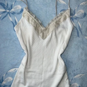 Vit ribbad linnetopp med spets Hollister - Supersöt vit ribbad linnetopp från Hollister med tunna axelband och v-ringad spetskant framtill. Materialet är mjuk bomull med lite stretch och spetsdetaljer vid halsen. Perfekt för dig som gillar en clean och feminin stil.