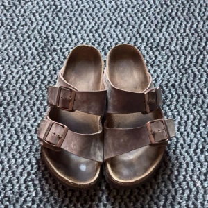 Bruna Birkenstock sandaler i skinn - Säljer ett par klassiska bruna Birkenstock sandaler med två breda remmar och justerbara metallspännen. Ovandelen är i skinn och sulan är ergonomiskt formad för extra komfort. Perfekta för sommaren och riktigt sköna att gå i.