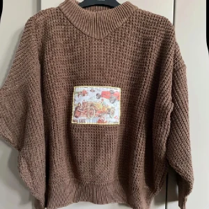 Brun stickad tröja med bilpatch  - Säljer en brun, grovstickad tröja med lös passform och hög krage. På framsidan finns en unik patch med vintage bilmotiv i färgerna röd, gul och blå. Tröjan har långa ärmar och breda muddar. Perfekt för dig som gillar retrodetaljer och vill sticka ut.