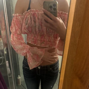 Blommig offshoulder croppad topp - Säljer en superfin blommig offshoulder topp i rosa och ljusa toner. Toppen är croppad med smockad midja och vida, långa ärmar. Tunt och luftigt material, perfekt för varma dagar. Romantisk vibe och snygg passform. Säljer då den är för liten.