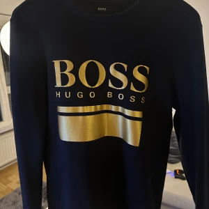 sweatshirt från Hugo Boss med guldtryck - Navyblå  sweatshirt från Hugo Boss med stort guldigt BOSS-tryck och bred guldfärgad detalj framtill. Tröjan har rund hals, långa ärmar och ribbade muddar. Perfekt för dig som gillar stilrena och lyxiga streetwear-plagg.