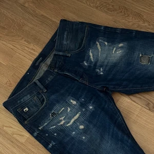 Jeans med slitningar och randig back ficka - Säljer ett par blå jeans från Scotch & Soda med coola slitningar och lagningar framtill. Baksidan har en unik bakficka med vitt och blått randigt tyg. Jeansen har smal passform och klassisk femficksdesign. Perfekta för dig som gillar en edgy streetstil.