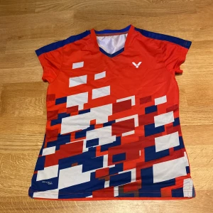 Victor - Röd sportig t-shirt - Badminton  - Snygg röd t-shirt från Victor med digitalt mönster i vitt och blått. T-shirten har v-ringning, korta ärmar och är tillverkad i ett lätt och svalt polyestermaterial. Perfekt för dig som gillar sportig stil och vill sticka ut på träningen.