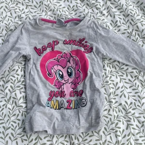Grå My Little Pony långärmad tröja - Superfin grå långärmad tröja med tryck av Pinkie Pie från My Little Pony och texten 'keep smiling you are amazing' i färgglada bokstäver. Rosa detaljer runt halsen och stort rosa hjärta på bröstet. Perfekt för dig som älskar söta motiv och färg!