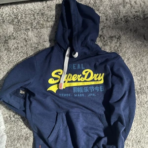 Superdry Hoodie - Säljer en blå hoodie från Superdry med gul och blå text på framsidan. Tröjan har en justerbar huva med vita snören och en stor ficka framtill. Perfekt för en avslappnad stil.