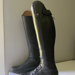 Svarta ridstövlar från Ariat, strl 38 - Snygga svarta ridstövlar från Ariat i äkta skinn med snörning framtill och dragkedja bak. Modellen är slim i vaden och har en klassisk, hög siluett med diskreta detaljer upptill. Perfekta för dig som vill ha en stilren look i stallet.