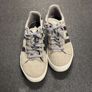 Grå sneakers från Adidas, strl 42 2/3 - Snygga grå sneakers från Adidas med klassiska svarta ränder på sidorna och vit sula. Skorna har snörning och är tillverkade i textilmaterial. Perfekta för dig som gillar en clean och stilren look till vardags. Inköpta 2023 i USA, sparsamt använda pga fel storlek.