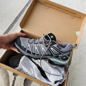 Salomon grå sneakers strl 39 1/3 - Säljer ett par grå och svarta Salomon sneakers med coola detaljer i mintgrönt och silver. Skorna har snörning, robust sula och mesh-överdel för extra komfort. Perfekta för dig som gillar en sportig och modern look. Storlek 39 1/3 (EU). Ord.pris 1600kr, helt nya