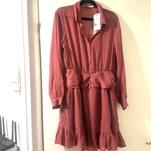 Rostrosa kortklänning från NA-KD aldrig använt - Supersöt rostrosa kortklänning från NA-KD med markerad midja, volangkant nertill och diskret strukturmönster. Klänningen har långa ärmar, knappar framtill och klassisk krage. Perfekt för dig som gillar en feminin och trendig look.