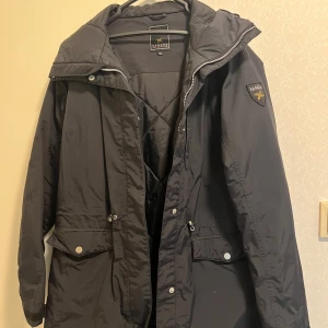 Svart parkajacka från Canard Collection - Svart parkajacka från Canard Collection med huva, dragkedja och knappar framtill. Jackan har två stora fickor med lock och snyggt broderat märke på ärmen. Materialet är slitstark polyester och modellen är normal i passningen. Perfekt för kyliga dagar.