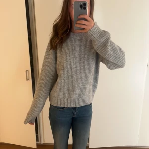 Grå stickad tröja - Mysig grå stickad tröja med rund halsringning och långa ärmar. Tröjan har en ribbad kant vid hals, ärmslut och nederkant. Perfekt för lager-på-lager och chill dagar. Enkel och stilren design som passar till jeans eller kjol.