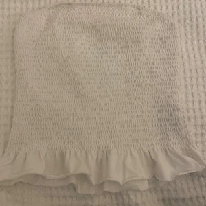 Vit bandeau tubtopp med volang från Vila - Supersöt vit bandeau tubtopp från Vila med smockad struktur och volangkant nertill. Toppen är ärmlös och har en mjuk, stretchig känsla som sitter skönt på kroppen. Perfekt med en snygg kofta över💕