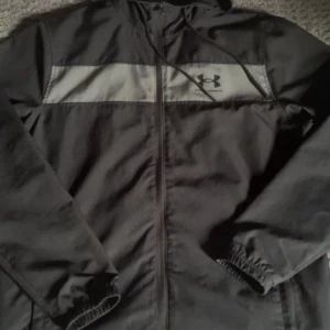 Under Armour vindjacka - Säljer en svart under armour vindjacka i fint skick. Den är sparsamt använd och har inga tecken på användning. Storleken är S men funkar även på M. Hör av dig vid frågor eller funderingar!