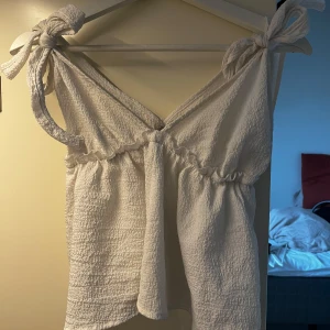 Vit linnetopp med knytaxlar - Supersöt vit linnetopp med knytbara axelband och lätt rynkad söm under bysten. Toppen har en luftig och flowy passform med struktur i tyget som ger en trendig känsla. Perfekt för varma dagar och enkel att styla med jeans eller kjol.