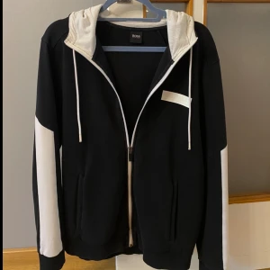 Hugo boss hoodie svart vit - Snygg svart och vit hoodie från Hugo Boss. Tröjan har dragkedja, huva med vita snören och vita paneler längs ärmarna. Perfekt för en chill och stilren look. Tillverkad i mjuk bomullsmix för extra komfort. Nyköpt i England för över 2000kr, går ej att få tag på den modellen längre, men den är tyvärr lite för stor för mig. Storlek M känns som M/L . Pris går att diskutera 