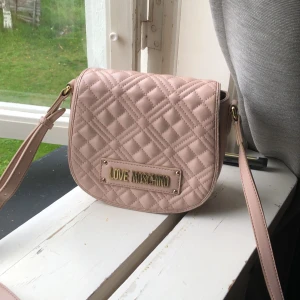 Rosa axelväska Love Moschino - Supersnygg rosa axelväska från Love Moschino med quiltad framsida och guldfärgade detaljer. Väskan har justerbar axelrem med logga, magnetlås och innerfoder med tryckt mönster. Perfekt storlek för det viktigaste och ger en lyxig vibe till din outfit.