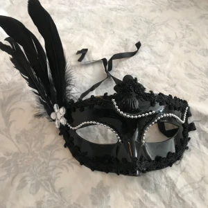 Svart maskeradmask med fjädrar - Snygg svart maskeradmask med glansig finish, dekorerad med svarta fjädrar på sidan och silvriga pärlor längs kanterna. Masken har svarta band för att knyta runt huvudet och en blomformad detalj med stenar vid fjädrarna. Perfekt för maskerad eller temafest.