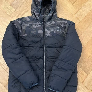 Svart och grå camo pufferjacka NYC - Snygg pufferjacka från NYC med svart och grått camouflagemönster. Jackan har huva, hel dragkedja och quiltad design. Perfekt för kalla dagar och har en cool streetstil. Vändbar modell med helsvart insida och camo på utsidan.