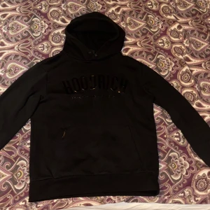 Svart hoodie från Hoodrich - Svart hoodie från Hoodrich med stor logga framtill. Klassisk känguruficka och huva med snörning. Perfekt för dig som gillar streetwear och vill ha en stilren look. Mjuk och skön, passar till många outfits.