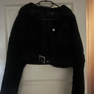 Svart croppad teddyjacka med bälte - Säljer en svart croppad teddyjacka med lång ärm och fint bälte i midjan. Jackan har en fluffig textur och stängs med knappar framtill. Jackan har även knappar vid ärmen men vissa knappar är lösa och håller inte riktigt så dem knäpps upp men det går att fixa 😊