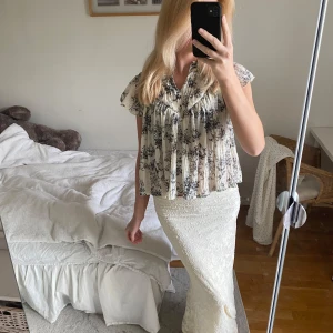 Blommig plisserad blus i vitt och svart - Säljer en luftig och flowy blus med svart blommönster på vit botten. Blusen har korta ärmar, v-ringning och plisserad design som ger en avslappnad och söt look. Perfekt för vår och sommar, lätt att matcha med både kjol och jeans.