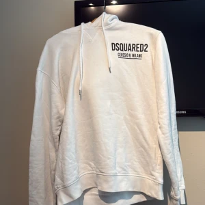 Vit hoodie från Dsquared2 - Säljer en vit hoodie från Dsquared2 med svart tryck på bröstet och snörning i huvan. Klassisk passform med långa ärmar och ribbade muddar. Perfekt för dig som gillar stilrena och exklusiva plagg.