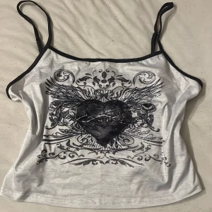 Ljus grått  linne med svart hjärtmotiv - Säljer ett ljus grått  linne med tunna svarta axelband och ett stort svart hjärta med vingar och detaljerad grafisk design framtill. Perfekt för dig som gillar y2k  och unik stil.