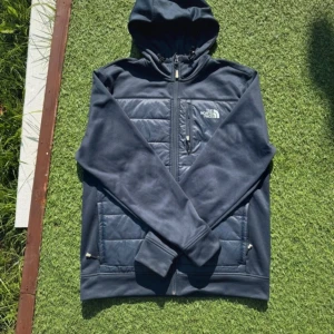 Mörkblå cardigan från The North Face - Säljer en mörkblå hoodie från The North Face med dragkedja och huva. Tröjan har quiltade partier framtill, bröstficka med dragkedja och två sidofickor. Perfekt för kyliga dagar och outdoor-häng.