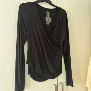 Svart omlottopp från Kappahl - Svart omlottopp från Kappahl med lång ärm och djup v-ringning. Tunn och mjuk i materialet, perfekt för en stilren look. Snygg drapering framtill som ger en cool touch. Tröjan är omlott