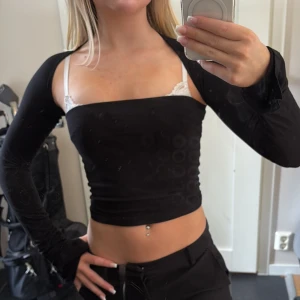Svart croppad bolerotopp - Säljer en svart croppad bolerotopp med långa ärmar. Toppen har en tight passform och är axelbandslös, perfekt för att skapa en trendig och edgy look. Materialet är stretchigt och bekvämt, vilket gör den enkel att styla till olika outfits.