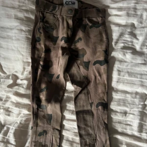 Camouflage ridbyxor från CRW, stl 160 - Säljer ett par bruna ridbyxor från CRW i coolt camouflage-mönster. Byxorna har klassisk passform med förstärkning på insidan av benen och midja med hällor. Perfekta för dig som gillar att sticka ut i stallet.