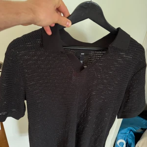 Svart stickad pikétröja från H&M - Aldrig använd - Säljer en svart stickad pikétröja från H&M med klassisk krage och kort ärm. Tröjan har ett luftigt, grovmaskigt material som ger en cool och avslappnad vibe. Perfekt för varmare dagar när du vill ha något stilrent men ändå bekvämt.