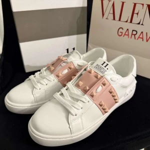 Valentino Rockstud sneakers vit/rosa - Snygga Valentino Garavani Rockstud sneakers i vitt skinn med ljusrosa band och ikoniska nitar. Klassisk rund tå, platt sula och snörning framtill. Perfekta för dig som vill ha en lyxig och trendig look med extra edge. VI HAR FRÅN STORLEK 37-39